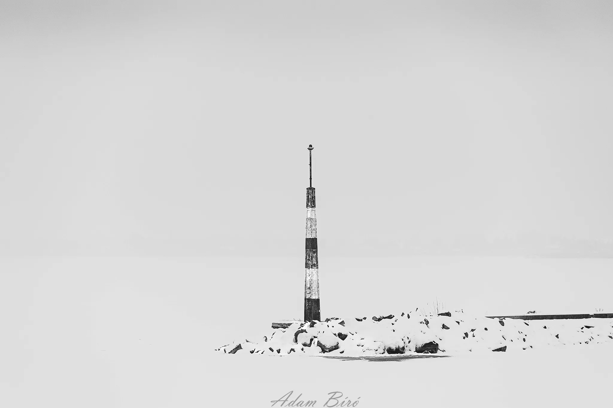 Minimalist black and white winter landscape at Lake Balaton with a solitary navigation marker emerging from snow and fog in Balatonföldvár, Hungary.