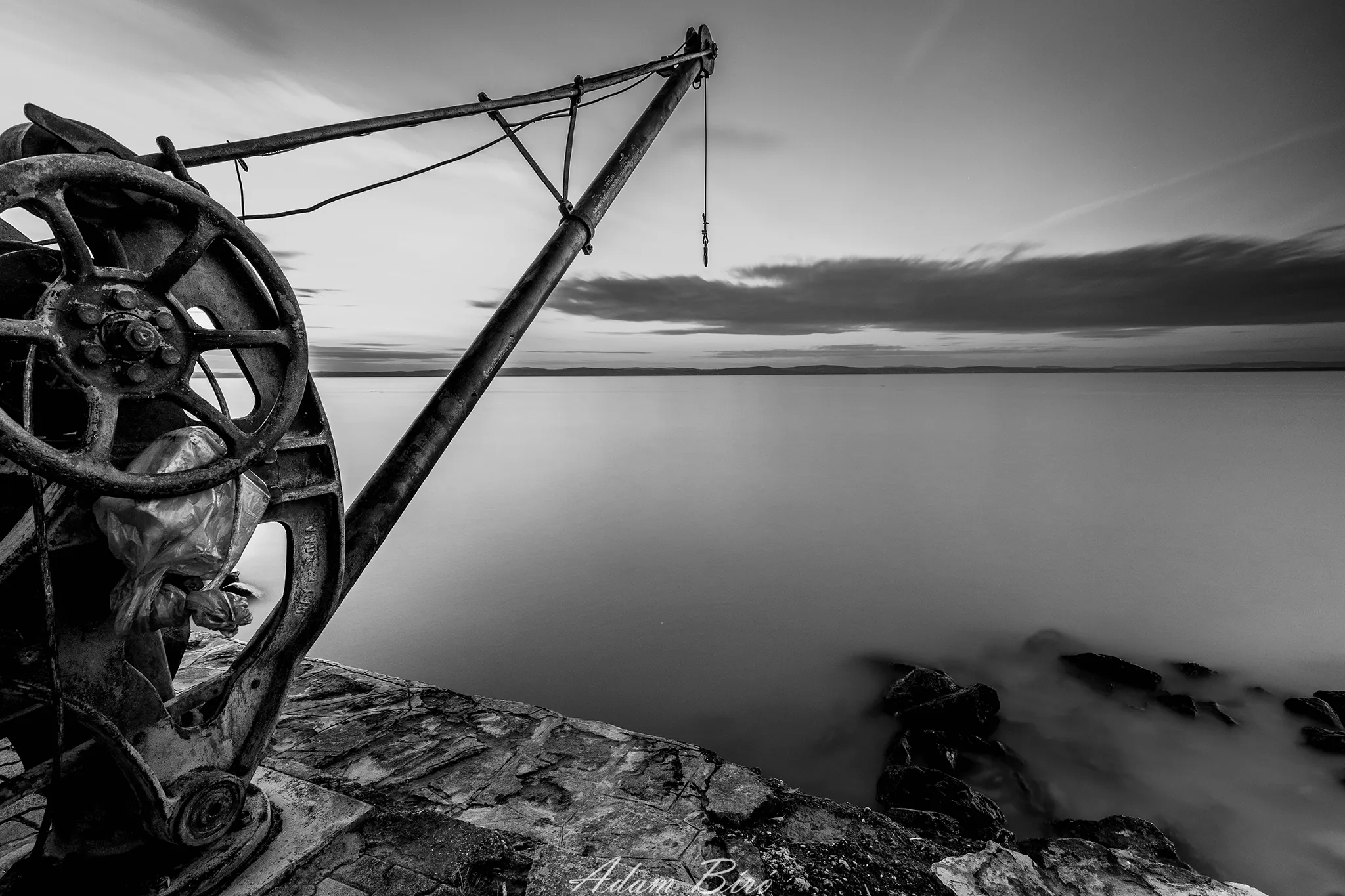 Black-and-white long exposure of an old crane by Lake Balaton extending over calm water with blurred rocks and distant horizon.