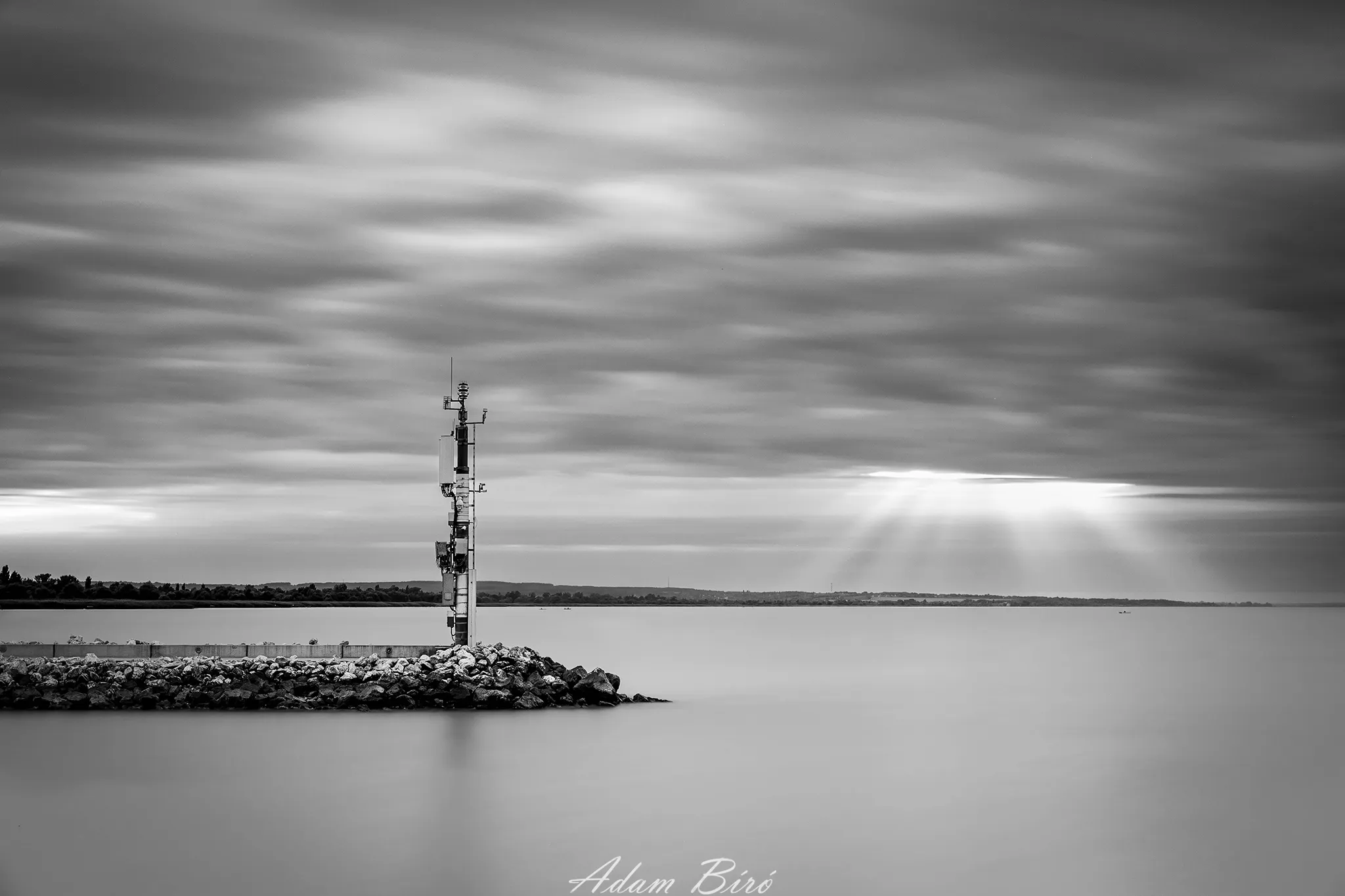 A solitary signal tower standing on rocks by Lake Balaton under dramatic clouds and light rays in a minimalist black and white scene.